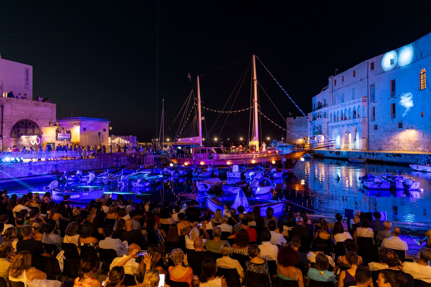 Il pubblico di Porto Rubino in riva al porto, di sera, tra barche e luci blu